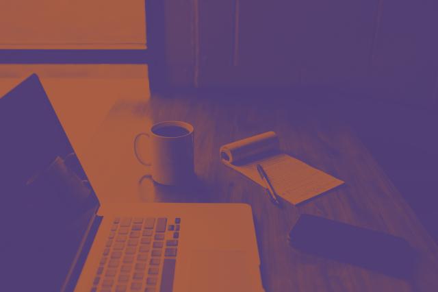 A phot of a desk with a laptop, cup of coffee and notebook